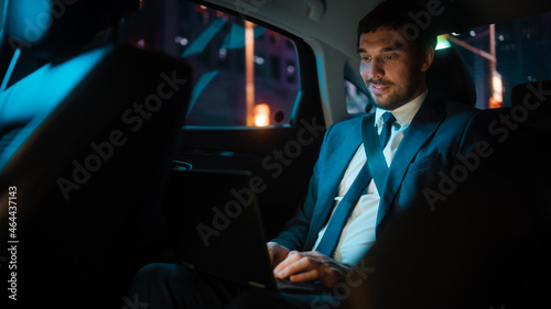 Handsome Businessman in a Suit Commuting from Office in a Backseat of His Luxury Car at Night. Entrepreneur Working on Laptop Computer while in Transfer Taxi in Urban City Street with Neon Signs. © Gorodenkoff