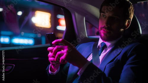 Successful Businessman in a Suit Commuting from Office in a Backseat of His Car at Night. Entrepreneur Using Smartphone while in Transfer Taxi in Urban City Street with Working Neon Signs. © Gorodenkoff