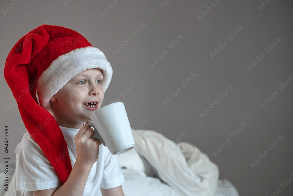 Portrait cute boy is sitting on the bed with a mug in a Santa hat. Happy holidays. Enjoying Christmas morning. space for text Portrait cute boy is sitting on the bed with a mug in a Santa hat. Happy holidays. Enjoying Christmas morning. space for text
