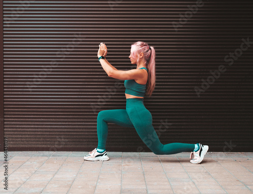 Fitness smiling woman in grey sports clothing with pink hair. Young beautiful model with perfect body.Female posing in the street near wall.Doing forward lunges. Stretching out before training