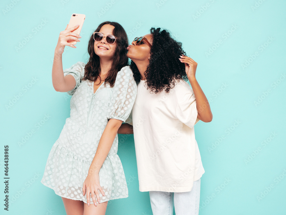 Two young beautiful smiling international hipster female in trendy summer clothes. Sexy carefree women posing near blue wall in studio. Positive models having fun in sunglasses. Taking selfie photos Two young beautiful smiling international hipster female in trendy summer clothes. Sexy carefree women posing near blue wall in studio. Positive models having fun in sunglasses. Taking selfie photos