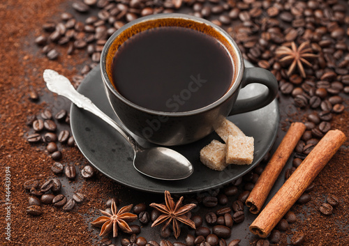 Black coffee in ceramic cup with cinnamon and cane sugar with anise star and silver spoon on ground coffee and beans.