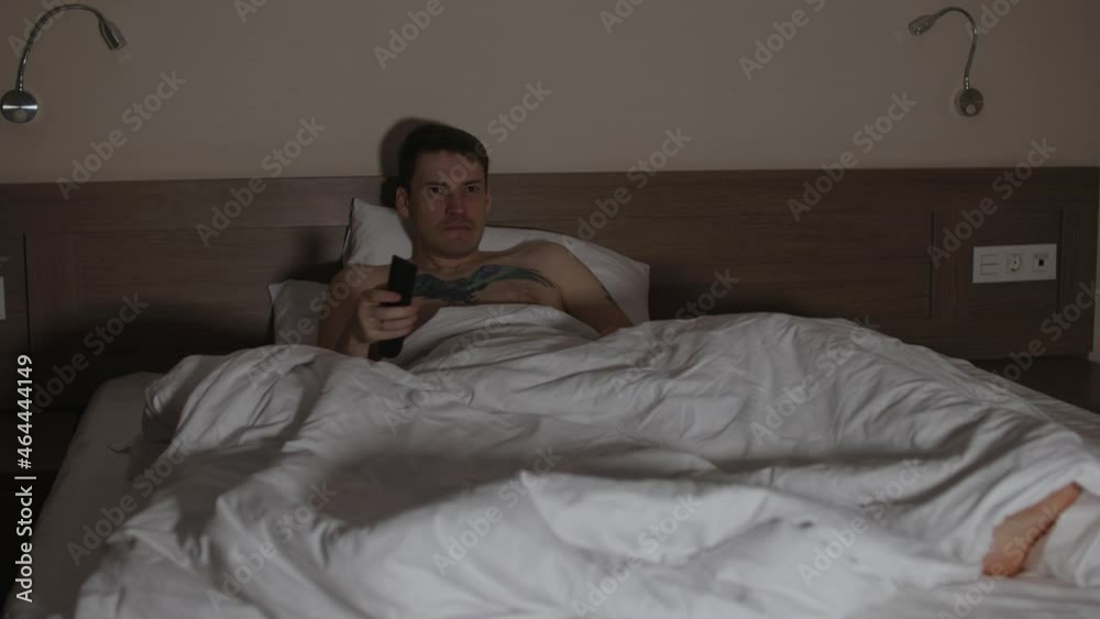 Young man lying on pillow under blanket in bed and watching tv in late evening. Relaxed male watching programs of show and switches channel with remote control in dark room.