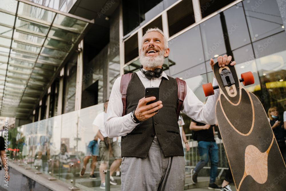 Grey mature man using mobile phone while walking with skateboard