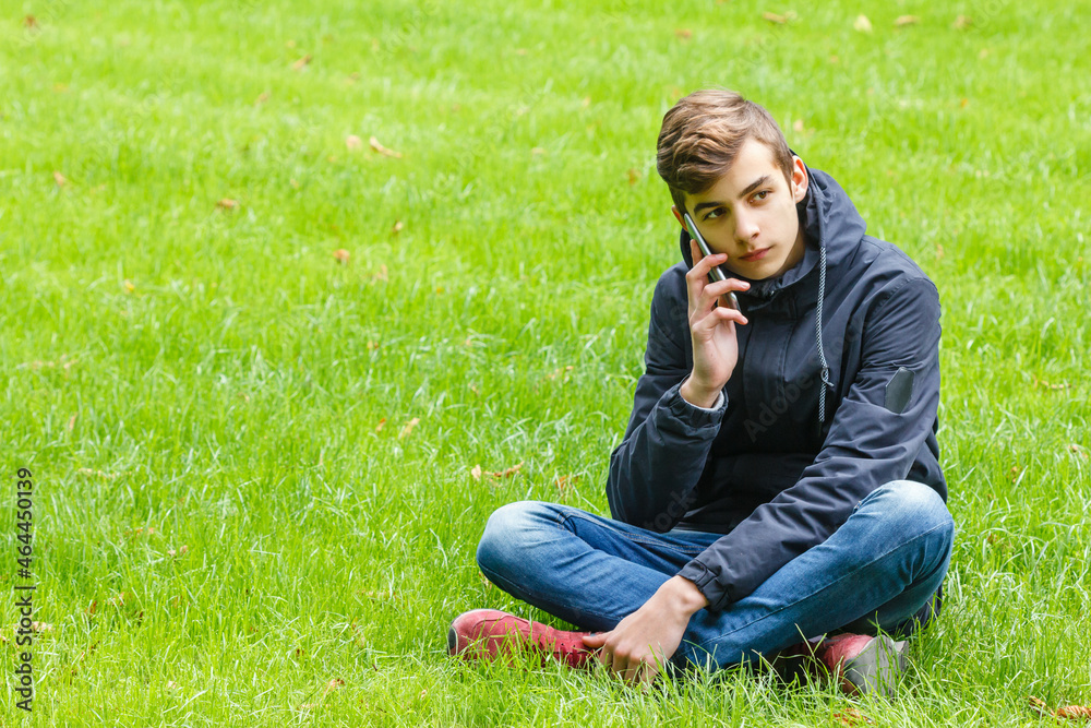 Male student talking on a phone seated on a grass in the city park Male student talking on a phone seated on a grass in the city park