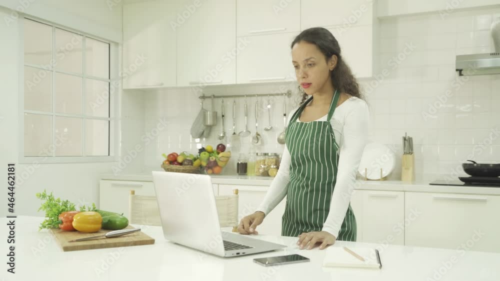 Portrait of happy smiling latin woman using a computer laptop notebook technology device, cooking food, working online at home in kitchen. People lifestyle. Household Portrait of happy smiling latin woman using a computer laptop notebook technology device, cooking food, working online at home in kitchen. People lifestyle. Household