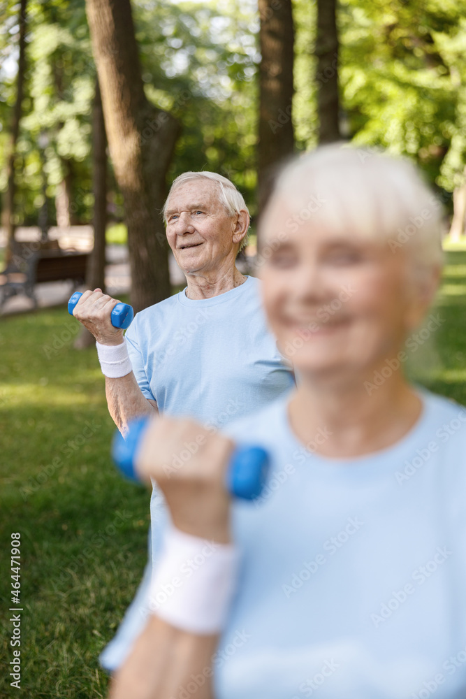 Happy senior gentleman does exercises with dumbbell training with wife in picturesque park
