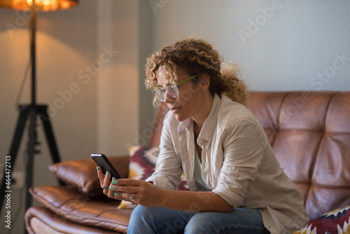 Serious young woman text messaging using mobile phone while sitting on sofa at home. Woman with curly hair and eyeglasses reading or texting using mobile phone in the living room