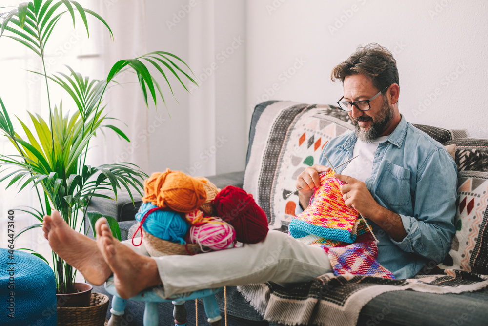 Mature man knitting wool with needle while sitting on sofa at home.  Caucasian man smiling and making sweater of wool while relaxing at home.  Woollen balls in basket on table. Photos