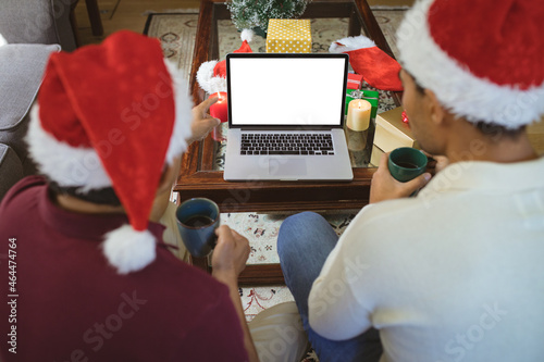 Biracial adult son and senior father in santa hats making laptop christmas video call