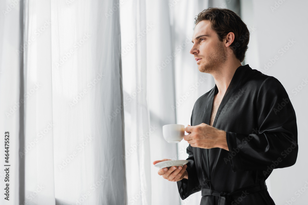 Young man in black robe holding coffee cup and saucer near curtains at home Young man in black robe holding coffee cup and saucer near curtains at home