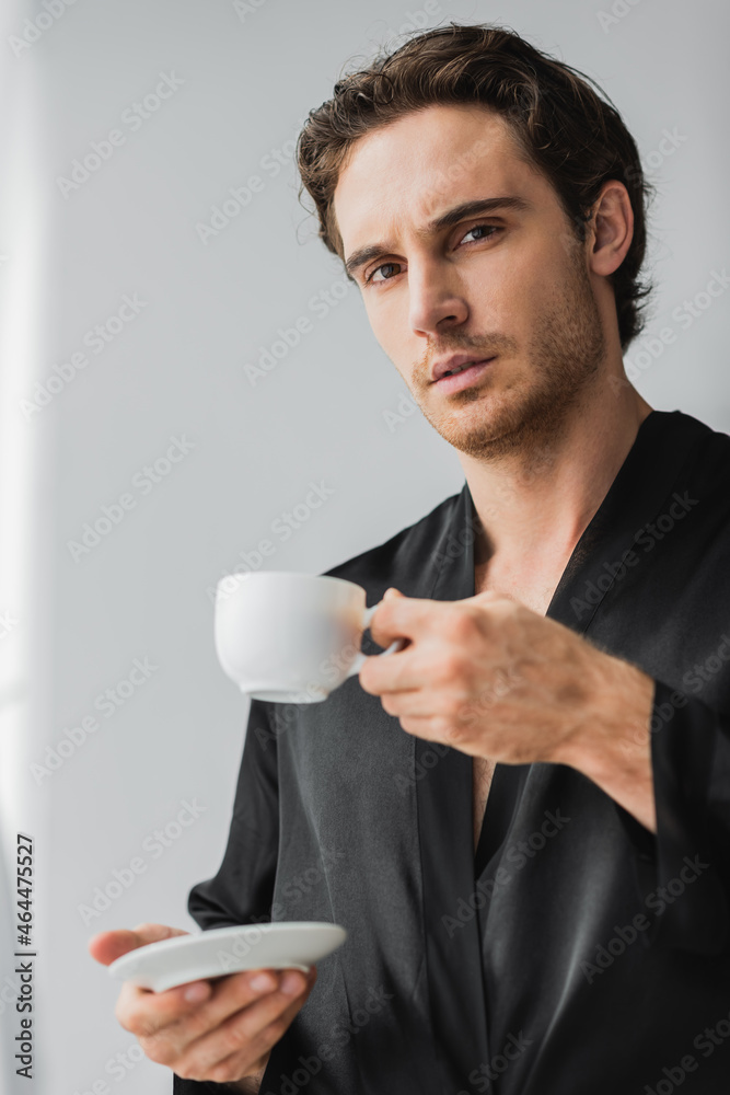 Young man in satin robe holding coffee cup with saucer and looking at camera at home Young man in satin robe holding coffee cup with saucer and looking at camera at home
