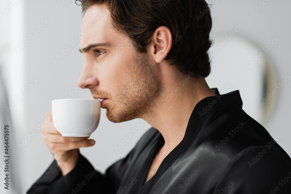 Side view of young man in robe holding coffee in bathroom Side view of young man in robe holding coffee in bathroom
