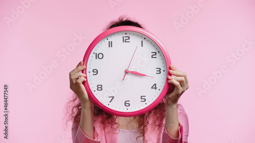 young woman obscuring face with wall clock isolated on pink