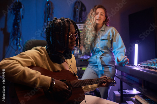 African young musician playing guitar while young woman singer in the background in the recording studio
