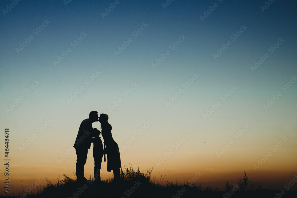 silhouette of a happy family with a child at sunset against the sky, parents hugging and kissing