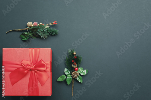 A red gift box with a bow and two ornate fir branches in the bottom corner against a dark emerald background. Top view. greeting card
