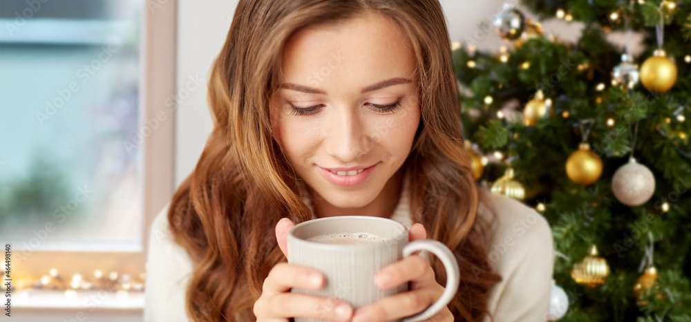 winter holidays, people and leisure concept - close up of smiling woman drinking hot chocolate at home over christmas tree lights background winter holidays, people and leisure concept - close up of smiling woman drinking hot chocolate at home over christmas tree lights background
