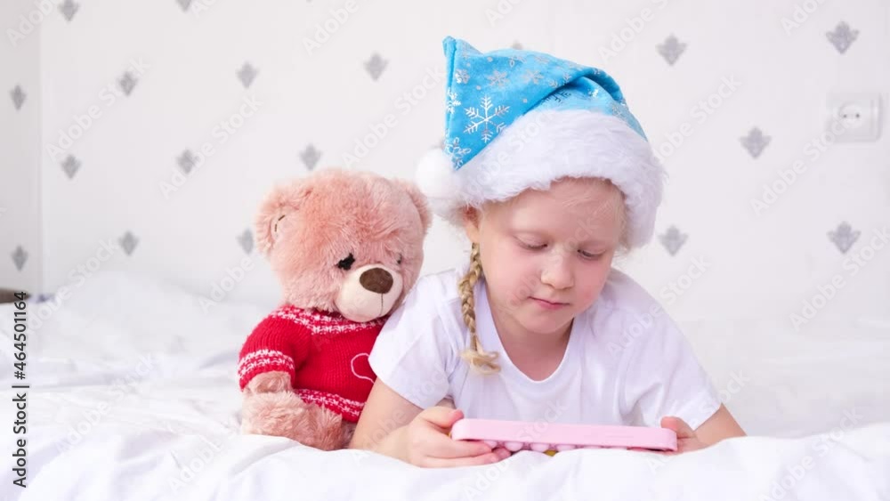 a girl in a blue santa claus hat looks at the screen a smart phone lies on the bed, a child watches a video on the phone a girl in a blue santa claus hat looks at the screen a smart phone lies on the bed, a child watches a video on the phone