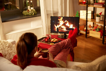christmas, winter holidays and leisure concept - young woman watching tv with fireplace on screen and drinking coffee with her feet on table at cozy home