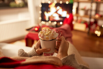 christmas, winter holidays and leisure concept - close up of young woman watching tv with fireplace on screen and holding mug of marshmallow and whipped cream resting feet on table at cozy home
