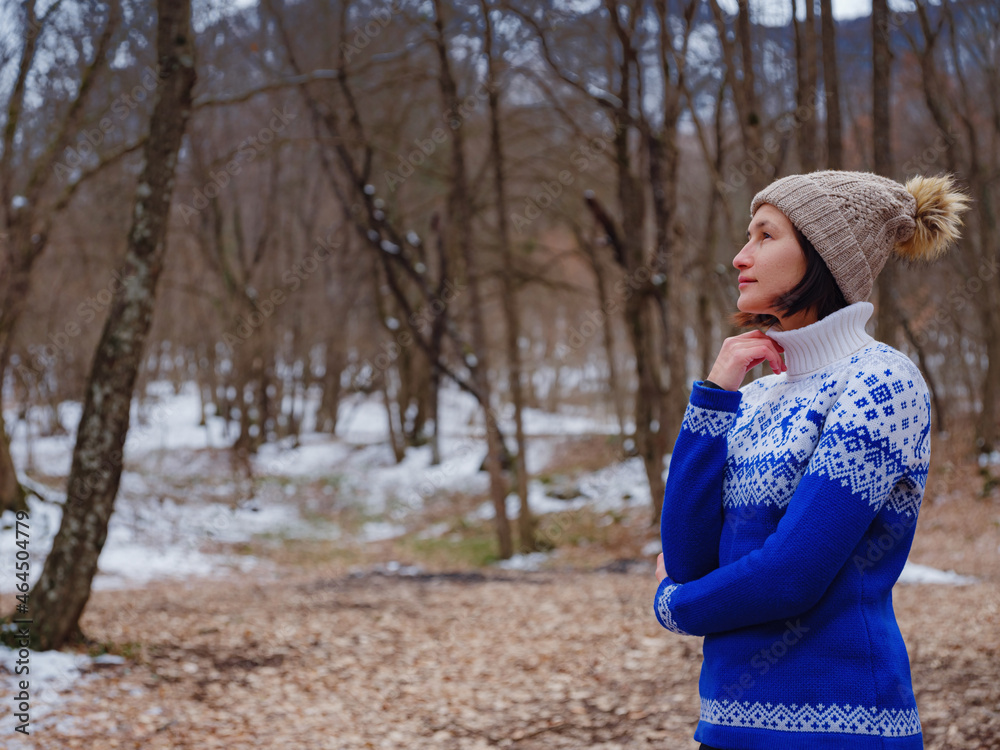 Beautiful woman standing among trees in winter forest. Wearing hat and blue sweater. Caucasian Asian female model outside in first snow