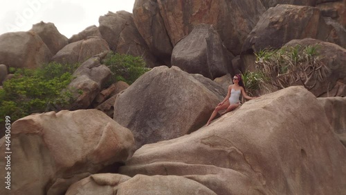 Young brunette woman with long hair in striped swimsuit and sunglasses poses sitting on huge rock on wild manless beach in Vietnam