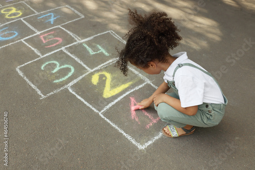 Little African American girl drawing hopscotch with chalk on asphalt outdoors. Happy childhood photo