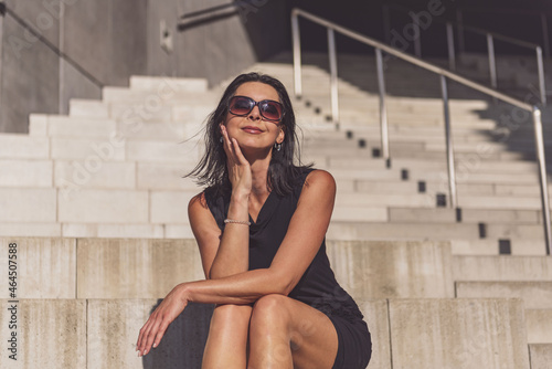 Dark-haired woman posing in a black dress on the concrete stairs. The woman is wearing black sunglasses. It's a beautiful sunny day.