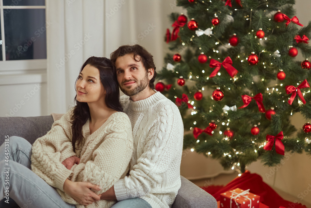 Marry Christmas Holidays and Happy New Year. Couple in love embracing on the sofa near the Christmas tree. Romantic tender couple in sweaters celebrating New Year at home near decorated fir tree and