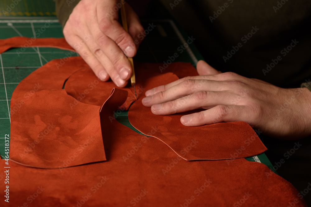 Shoemaker's hands holding craft knife cutting suede leather for handmade shoes on his workplace in workshop. Closeup.