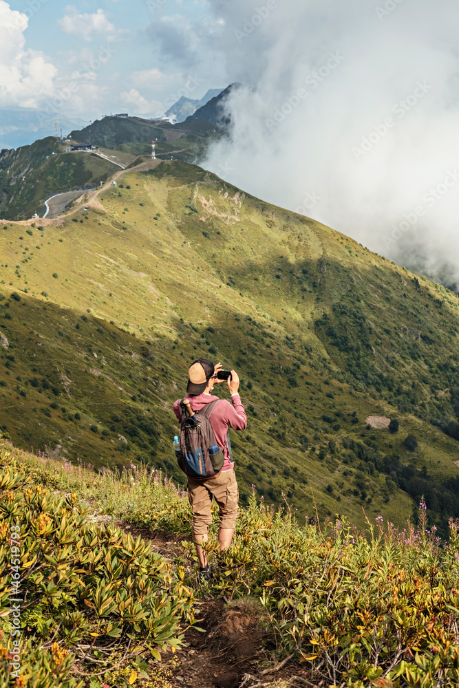 Young man with backpack hiking in Caucasus green mountains among bushes of rhododendron and taking photo on smartphone, using modern technology active and healthy lifestyle, landscape