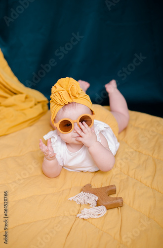 A fashionable baby girl is resting in yellow glasses. Cute girl posing in the studio in a yellow beanie cap