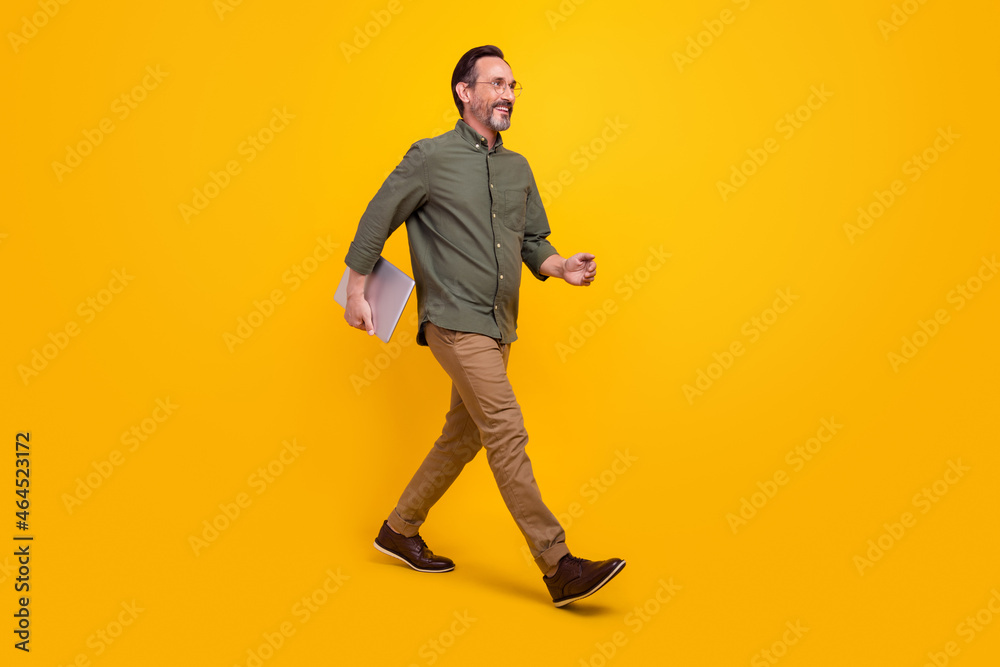 Full size profile side photo of mature business man happy positive smile go hold computer isolated over yellow color background Full size profile side photo of mature business man happy positive smile go hold computer isolated over yellow color background