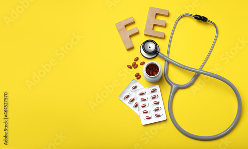 Wooden letters FE, pills and stethoscope on yellow background, flat lay with space for text. Anemia treatment