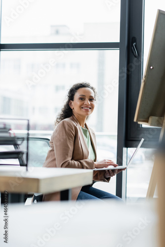 successful african american businesswoman smiling at camera while using laptop in office
