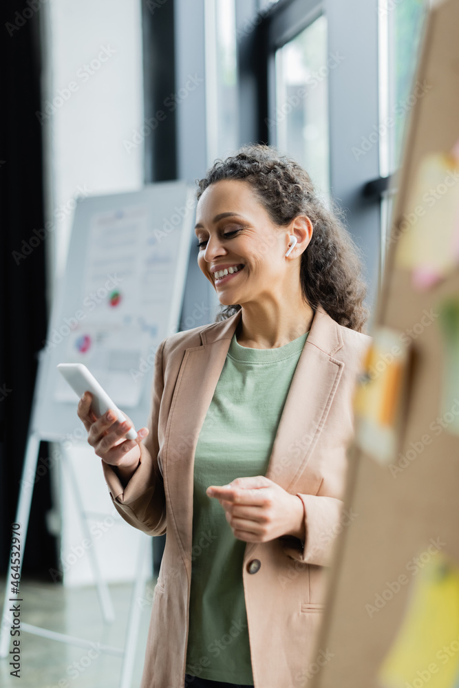 cheerful african american businesswoman in wireless earphone smiling during video call on smartphone on blurred foreground