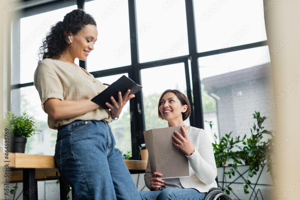 happy businesswoman in wheelchair holding documents near african american colleague writing in notebook happy businesswoman in wheelchair holding documents near african american colleague writing in notebook