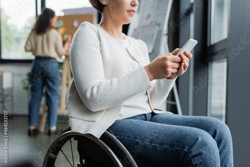 cropped view of businesswoman in wheelchair messaging on mobile phone near african american colleague on blurred background