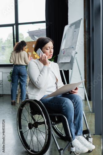 businesswoman in wheelchair looking at documents and talking on smartphone near blurred african american colleague