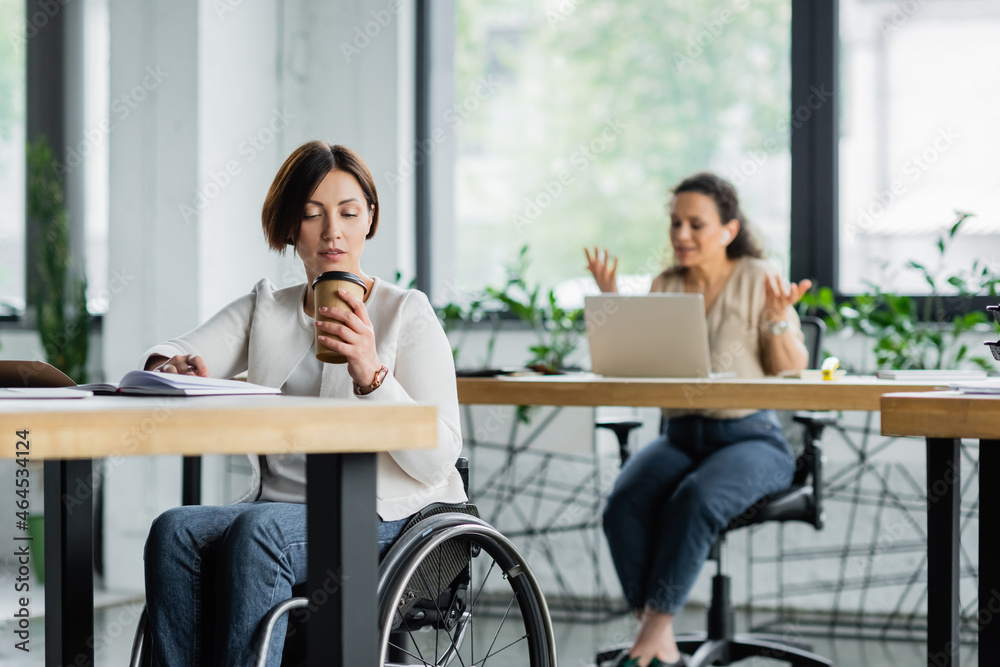businesswoman in wheelchair drinking coffee to go near african american colleague working on blurred background businesswoman in wheelchair drinking coffee to go near african american colleague working on blurred background