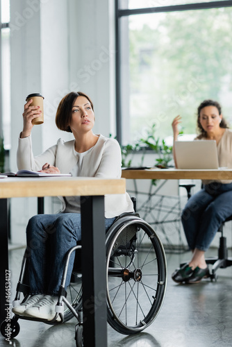 thoughtful businesswoman in wheelchair holding takeaway drink near blurred african american colleague in office