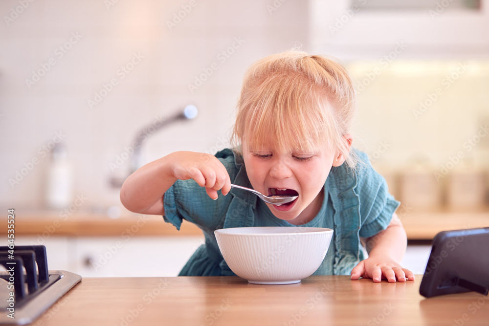 Young Girl At Home At Kitchen Counter Eating Breakfast Cereal From Bowl Whilst Watching Mobile Phone Young Girl At Home At Kitchen Counter Eating Breakfast Cereal From Bowl Whilst Watching Mobile Phone