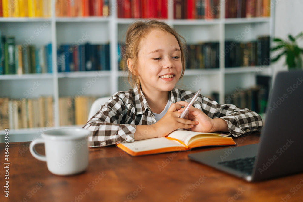 Close-up of cheerful pupil child school girl doing homework writing notes with pen in paper notebook and looking at laptop computer, selective focus. Concept of online e-learning education. Close-up of cheerful pupil child school girl doing homework writing notes with pen in paper notebook and looking at laptop computer, selective focus. Concept of online e-learning education.