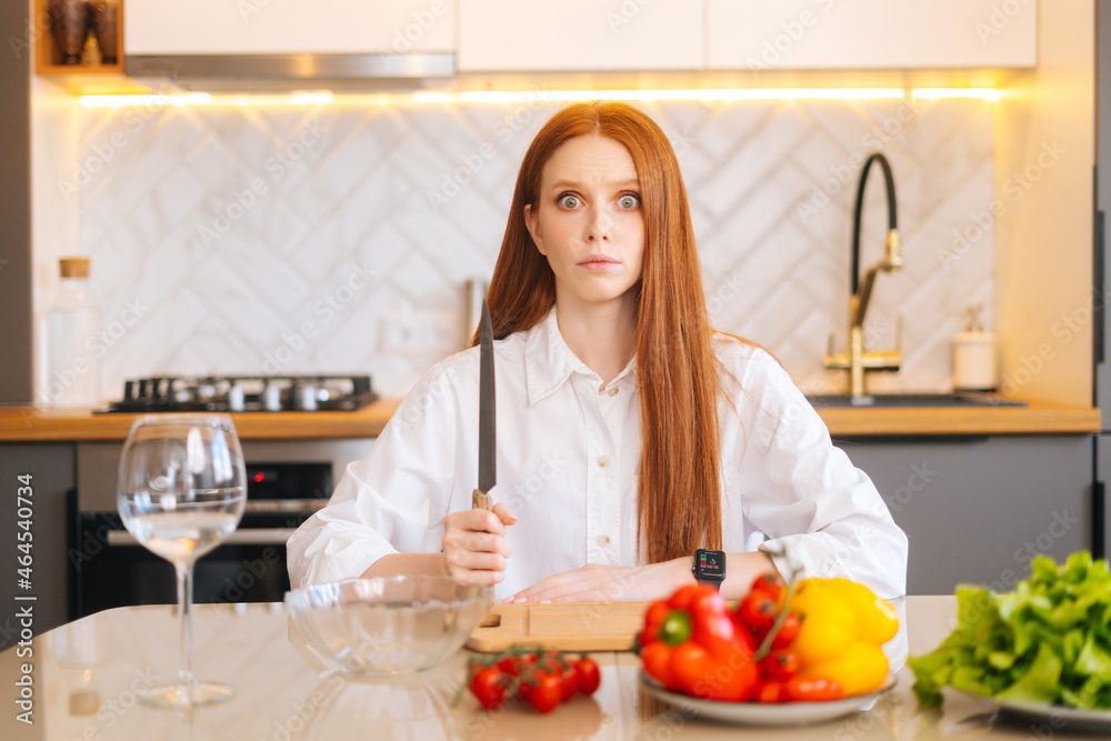 Portrait of attractive redhead young woman with crazy eyes holding big knife sitting at table with cutting board and looking at camera in light kitchen room with modern interior, close-up. Portrait of attractive redhead young woman with crazy eyes holding big knife sitting at table with cutting board and looking at camera in light kitchen room with modern interior, close-up.