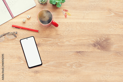 Mockup of a modern smartphone with on a wooden office desk table with notebook, glasses, plant, red cup of coffee and other office supplies. Top view, flat lay  with copy space photo