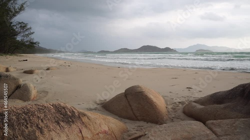 Small sea waves wash over sandy beach and rocks slowly under cloudy sky on quiet gloomy day with mountains on background in Vietnam