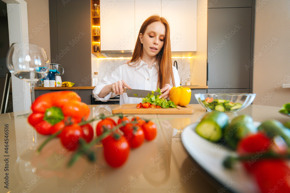 Front low-angle view of young redhead woman cutting fresh lettuce preparing food salad sitting at table in modern kitchen room. Pretty female cooking vegetarian dieting salad full of vitamins. Front low-angle view of young redhead woman cutting fresh lettuce preparing food salad sitting at table in modern kitchen room. Pretty female cooking vegetarian dieting salad full of vitamins.