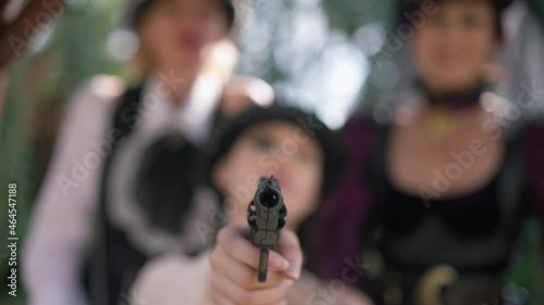 Close-up gun muzzle aiming at camera with three blurred steampunk women at background. Weapon gunpoint outdoors in slim female Caucasian hands. Slow motion photo