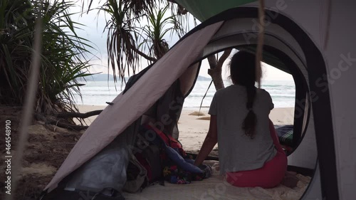Brunette girl with braid sits in small camp tent on tropical wild beach looking at waves in sea under cloudy sky in Vietnam